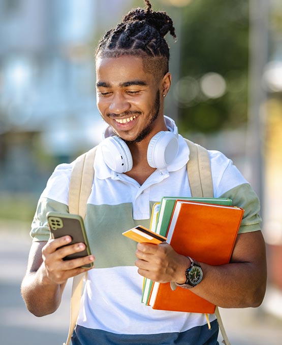 Male student holding books and a credit card looking at his phone.